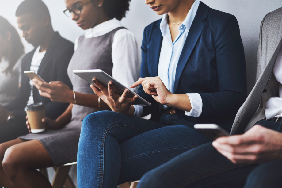 Business men and women sit against a wall working on tablets and mobiles