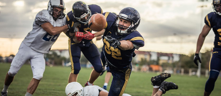 Football player dives for the ball while teammate blocks a tackle behind him.